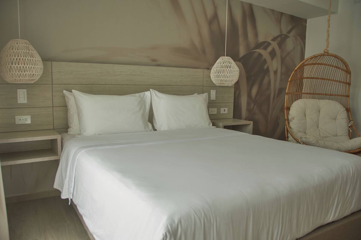 Un dormitorio con una cama grande, dos mesitas de noche, una silla colgante de mimbre y una lámpara fijada a la pared.