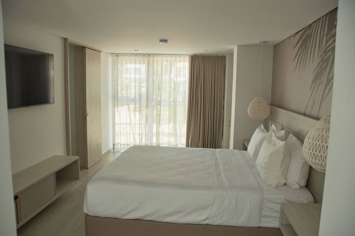 Habitación de hotel moderna con una cama grande, un televisor de pantalla plana, una mesita de noche y un ventanal con cortinas.