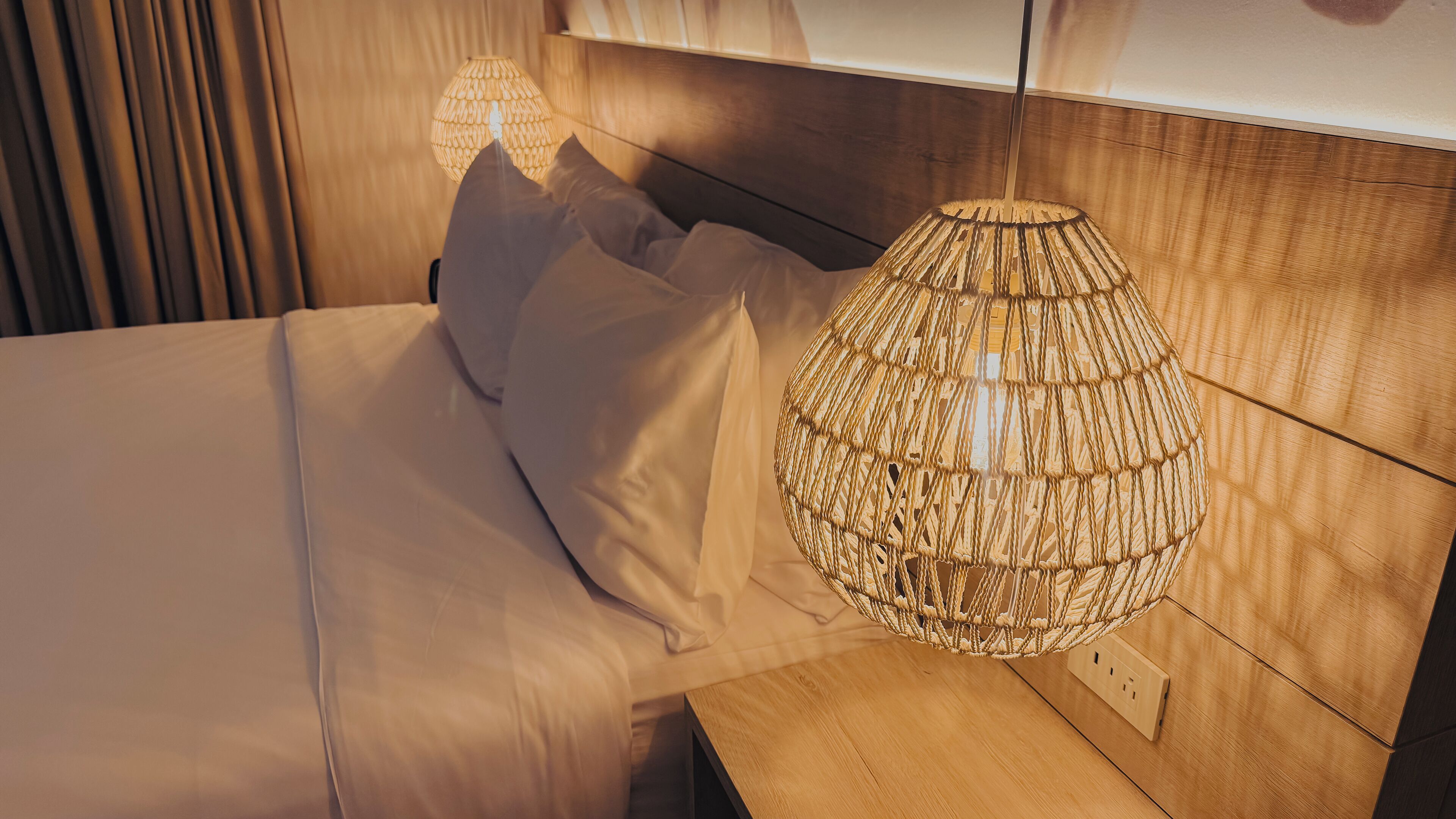 A neatly made bed with white linens and pillows, a wooden nightstand with a woven lamp, and a wooden headboard.