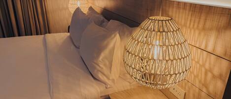 A neatly made bed with white linens and pillows, a wooden nightstand with a woven lamp, and a wooden headboard.