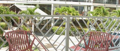 Un balcón con dos sillas rojas y una mesita pequeña, con vista a un edificio de varios pisos con balcones y vegetación.