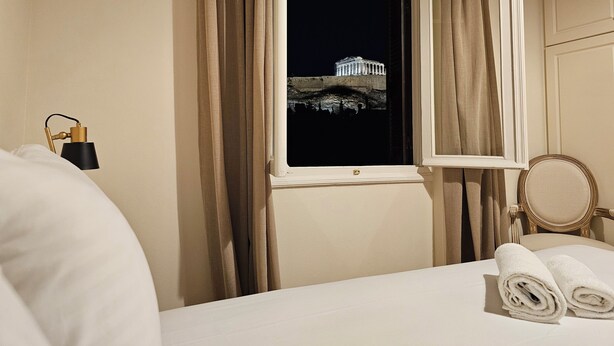 A hotel room with a bed, a nightstand with a lamp, a window with curtains, and a view of a building at night. - Acropolis Eternal Mansion by YHA  (Athens)