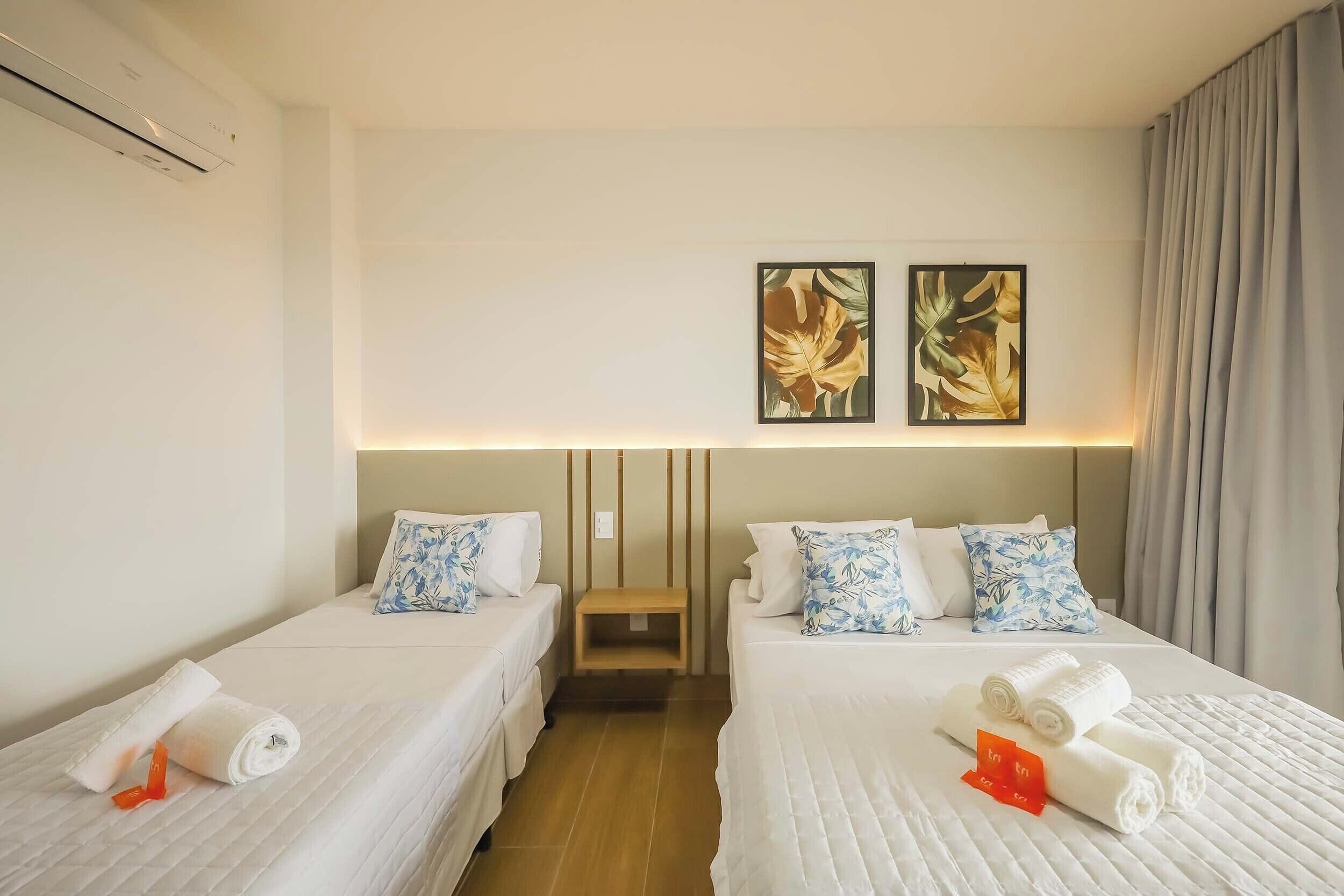 Une chambre d’hôtel avec deux lits, chacun équipé d’un oreiller et d’une serviette enroulée. Un climatiseur mural est fixé au mur, et deux tableaux encadrés sont accrochés au-dessus des lits.