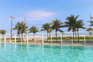 A clear swimming pool with a white railing, surrounded by palm trees and a view of the beach.
