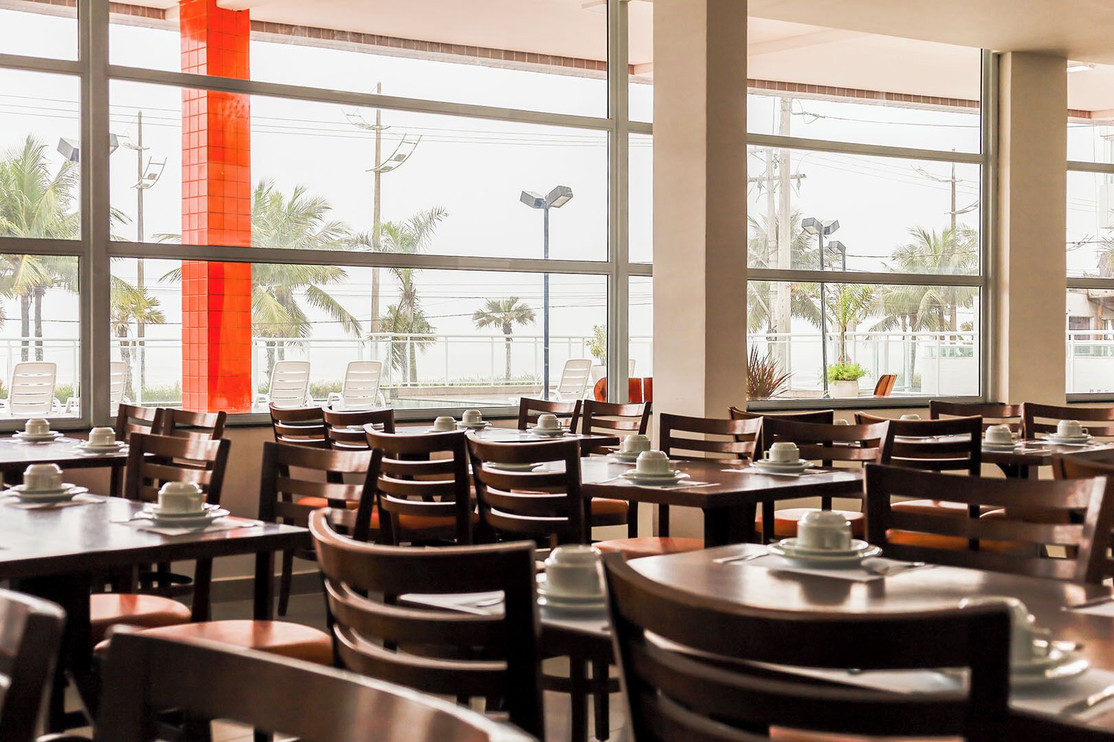 Un restaurante con varias mesas y sillas, amplios ventanales que ofrecen vistas a palmeras y edificios.