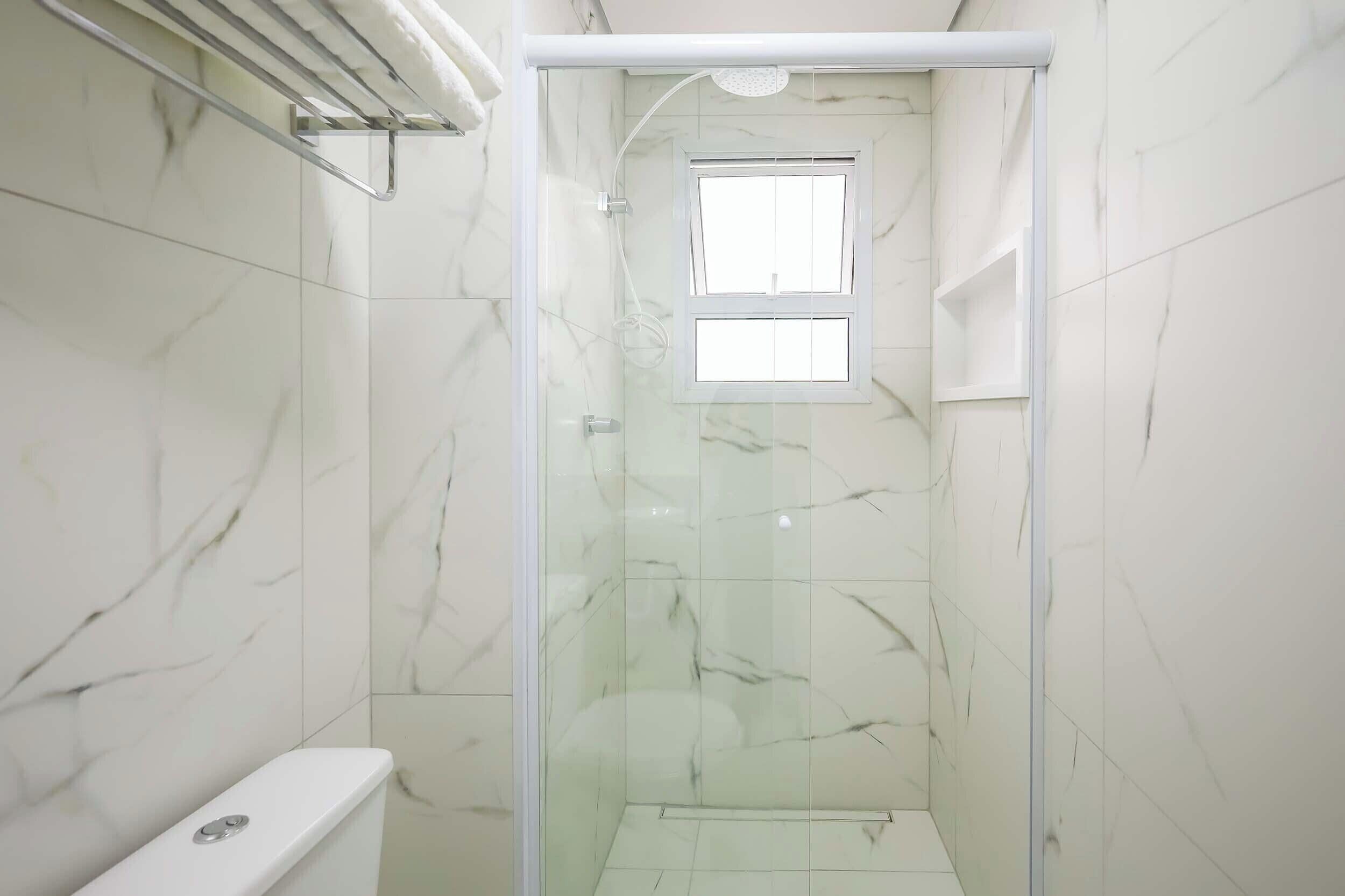 Un baño con cabina de ducha de vidrio, azulejos de mármol blanco y un ventanal.