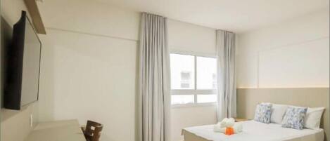 A hotel room with a bed, a TV mounted on the wall, a window with curtains, and a small table with a remote control.