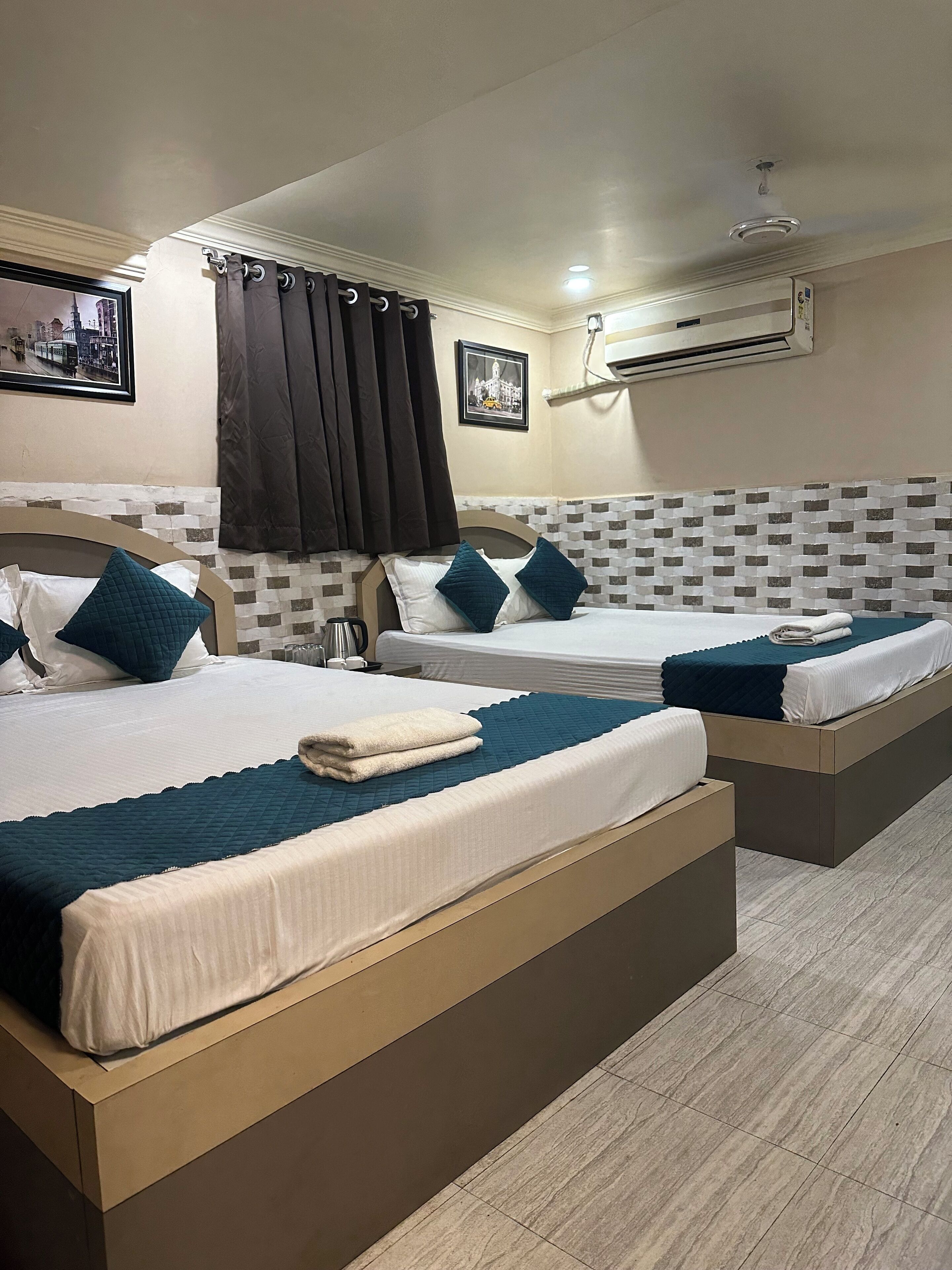 Une chambre d’hôtel avec deux lits, chacun recouvert d’une courtepointe bleue matelassée, avec du linge de lit blanc et des oreillers assortis de la même couleur. On y trouve une petite table avec une bouilloire et une tasse, ainsi qu’un tableau encadré accroché au mur.