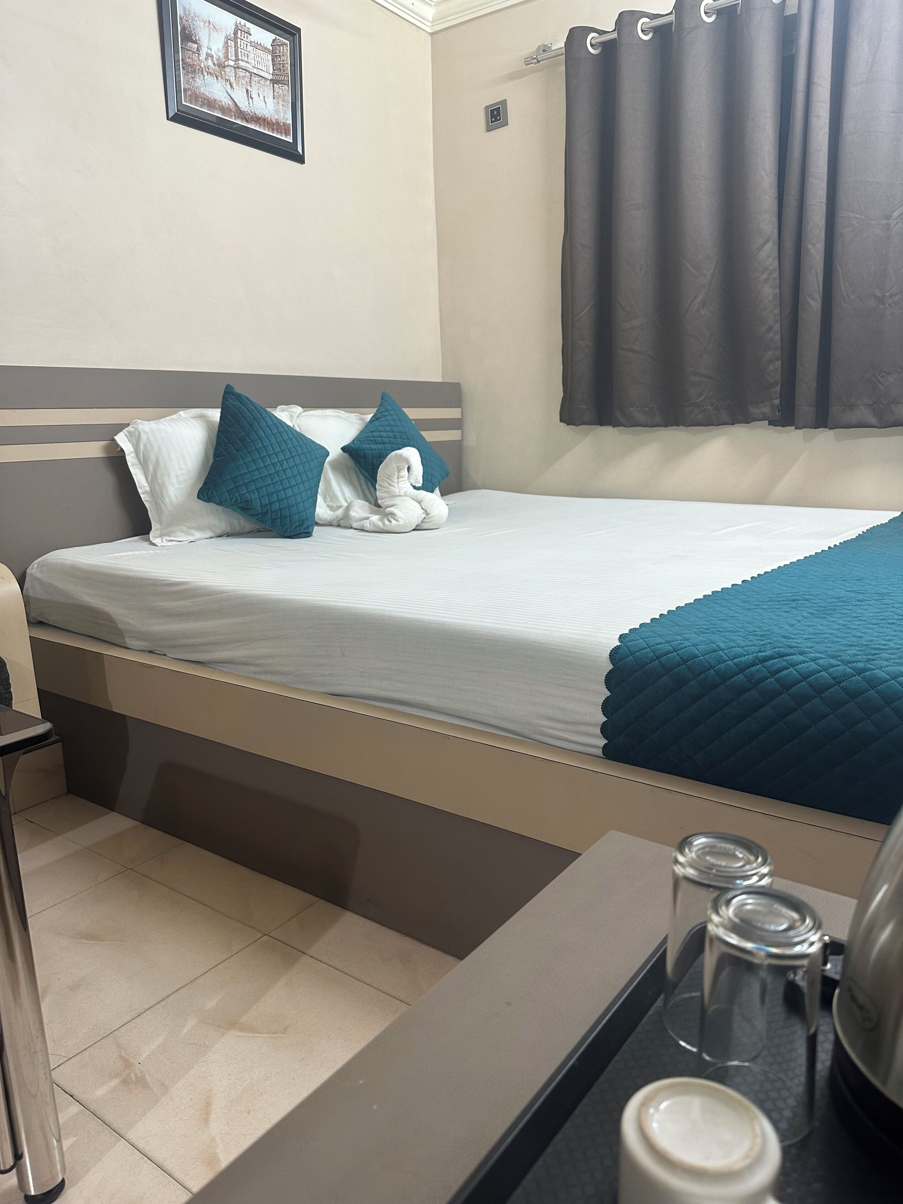 Cama bem arrumada com colcha branca e azul-turquesa, cortina escura e quadro na parede.