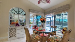 Uma sala de jantar com mesa de vidro, cadeiras de vime e vista para uma piscina e palmeiras.