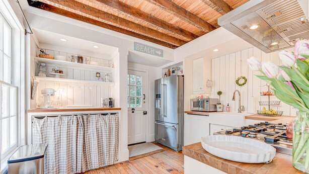 Une cuisine moderne avec un plancher en bois, des meubles blancs, un réfrigérateur en acier inoxydable et un panneau indiquant la direction de la plage.