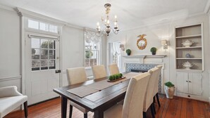 Une salle à manger avec une table en bois, des chaises et un lustre. On y trouve une fenêtre donnant sur l’extérieur, une cheminée et une étagère ornée d’objets décoratifs.