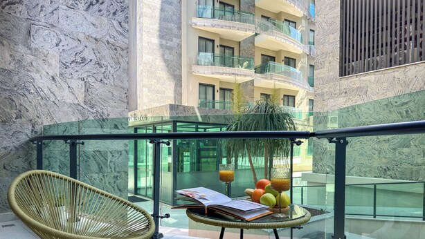 Un balcón con sillas de mimbre, una mesa de vidrio con frutero, y vista a un edificio con balcones.