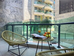 Un balcon avec des fauteuils en osier, une table en verre sur laquelle se trouve un panier de fruits, et une vue sur un immeuble doté de balcons.