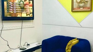 Un téléviseur fixé au mur affiche un menu, une chaise bleue avec un coussin jaune, et un tableau encadré représentant des fleurs.