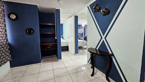 A hallway with a blue wall, a wooden shelf unit, a small table, and a white door leading to a bedroom with a bed and a nightstand. - Departamento Centro Escolar (Tehuacán)