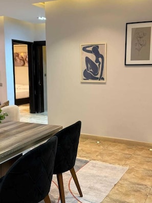 Un comedor con una mesa y sillas, una obra de arte enmarcada en la pared y una vista parcial de un dormitorio al fondo.