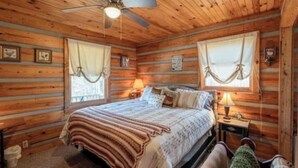 Une chambre à coucher dans une cabane en bois, avec un lit, deux lampes de chevet, un ventilateur de plafond et deux fenêtres avec des rideaux.
