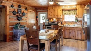L’intérieur d’une cabane en bois comprend une table à manger, des chaises, un coin cuisine et un ventilateur de plafond.