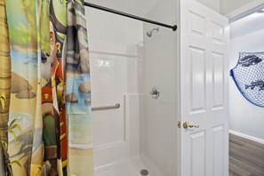 A shower area with a glass door, a showerhead, and a curtain with a cartoon character design. - The Pirate's Den Above Cedar Key Island Bakery (Cedar Key)