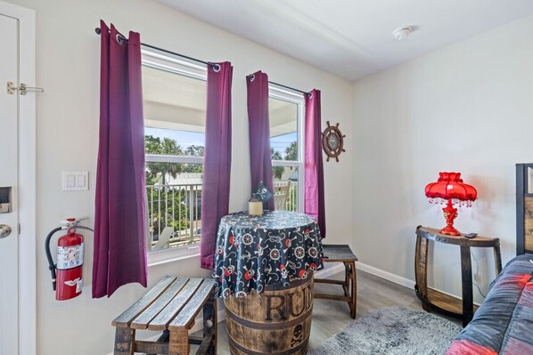 A room with a wooden barrel table, a small round table with a patterned tablecloth, a red lamp, and a view of greenery outside. - The Pirate's Den Above Cedar Key Island Bakery (Cedar Key)