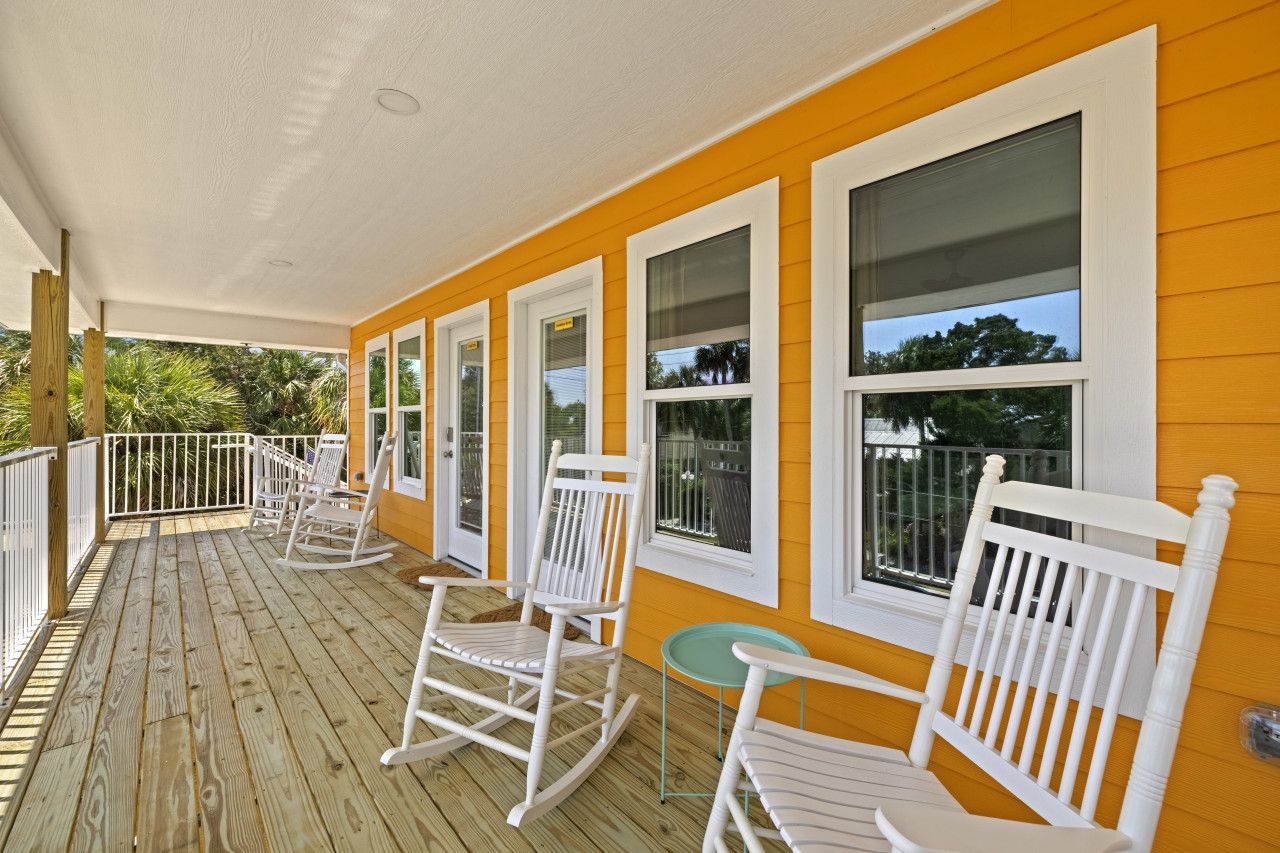 A covered porch with white rocking chairs and a small table, yellow siding, and large windows.