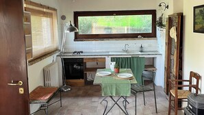 Une cuisine rustique avec un plafond en bois, une fenêtre donnant sur la verdure, un sol carrelé et un mobilier simple comprenant une table, des chaises et un poêle.