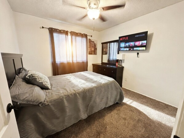 Un dormitorio con cama, cómoda, ventilador de techo, televisor montado en la pared y ventana con cortinas.