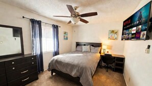 Un dormitorio con cama, escritorio, silla, ventilador de techo, televisor montado en la pared y cómoda.