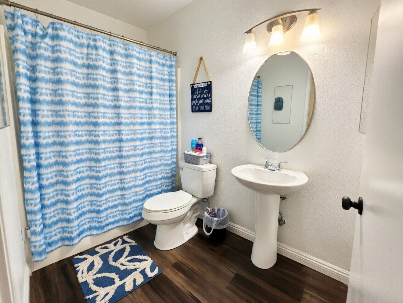 Une salle de bain avec des toilettes blanches, un lavabo blanc, un rideau de douche bleu et blanc, et un petit tapis.