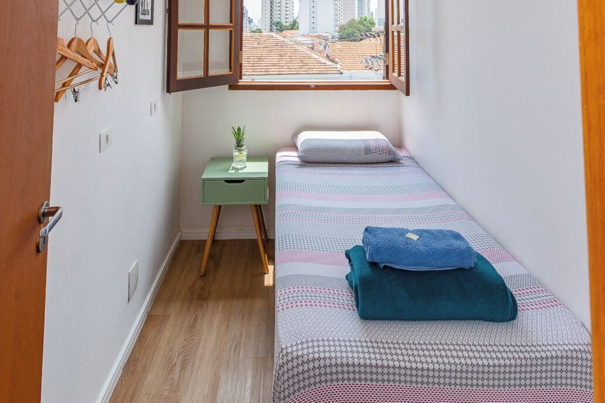 A small, bright room with a bed, a desk, and a window with an open view of buildings.
