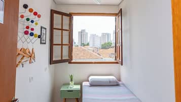 Um quarto pequeno e luminoso, com uma cama, uma escrivaninha e uma janela que oferece uma vista aberta para edifícios.