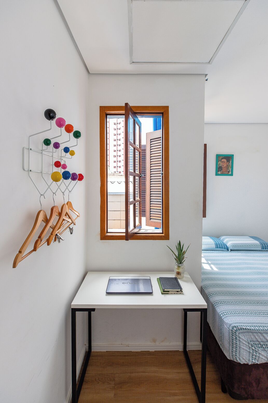 Una habitación con un escritorio blanco, una ventana con persianas de madera, un adorno esférico en la pared y una cama con cubrecama estampado.