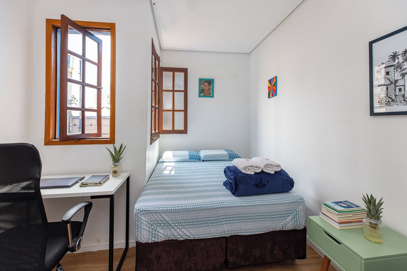 A small, well-lit bedroom with a bed, a desk, and a chair. There are two windows, one with a view of the outside.