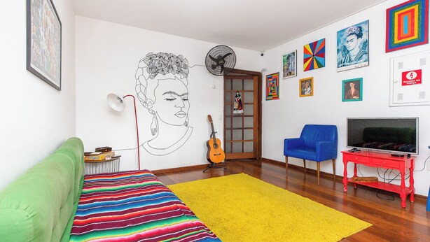 Una sala de estar con un sofá verde, una manta a rayas de colores, un tapete amarillo, una guitarra, una silla azul, un mueble rojo para televisor y diversas obras de arte en la pared.