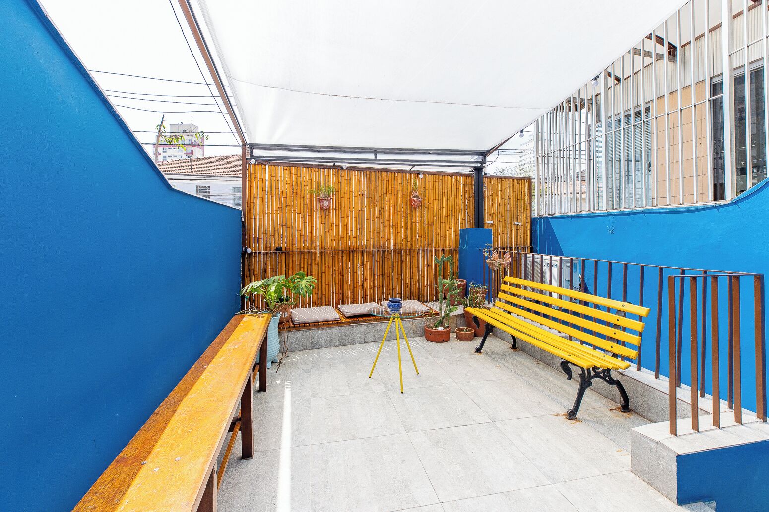A rooftop patio with a wooden bench, a yellow bench, and potted plants.