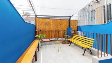A rooftop patio with a wooden bench, a yellow bench, and potted plants.