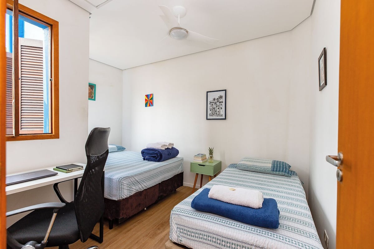 A small, tidy room with two beds, a desk, and a chair. There are two small side tables, one with a plant and a book, and the other with a lamp.