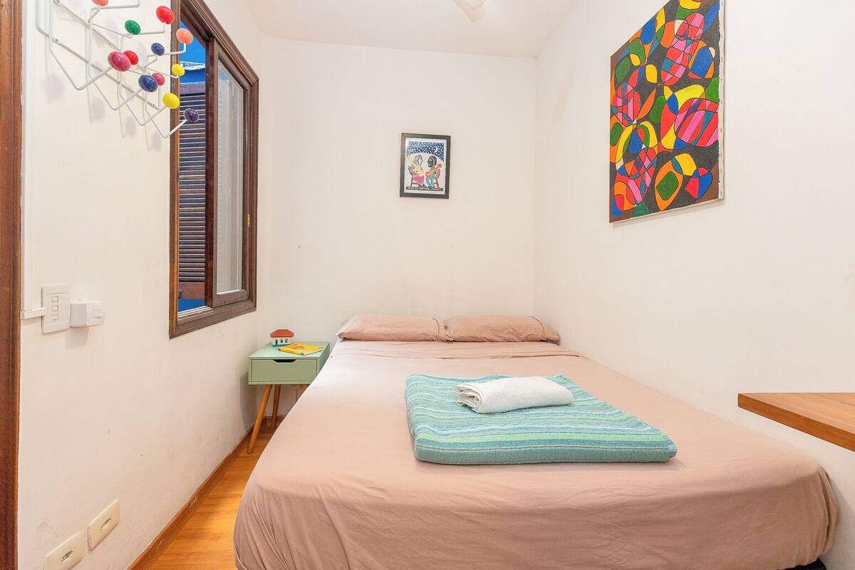 A small, neatly arranged bedroom with a single bed, a bedside table, a window with blinds, a wall-mounted artwork, and a colorful abstract painting.