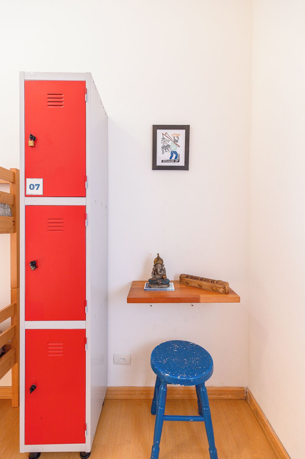 A room with a red locker, a blue stool, a wooden shelf with a statue and a book, and a framed picture on the wall.