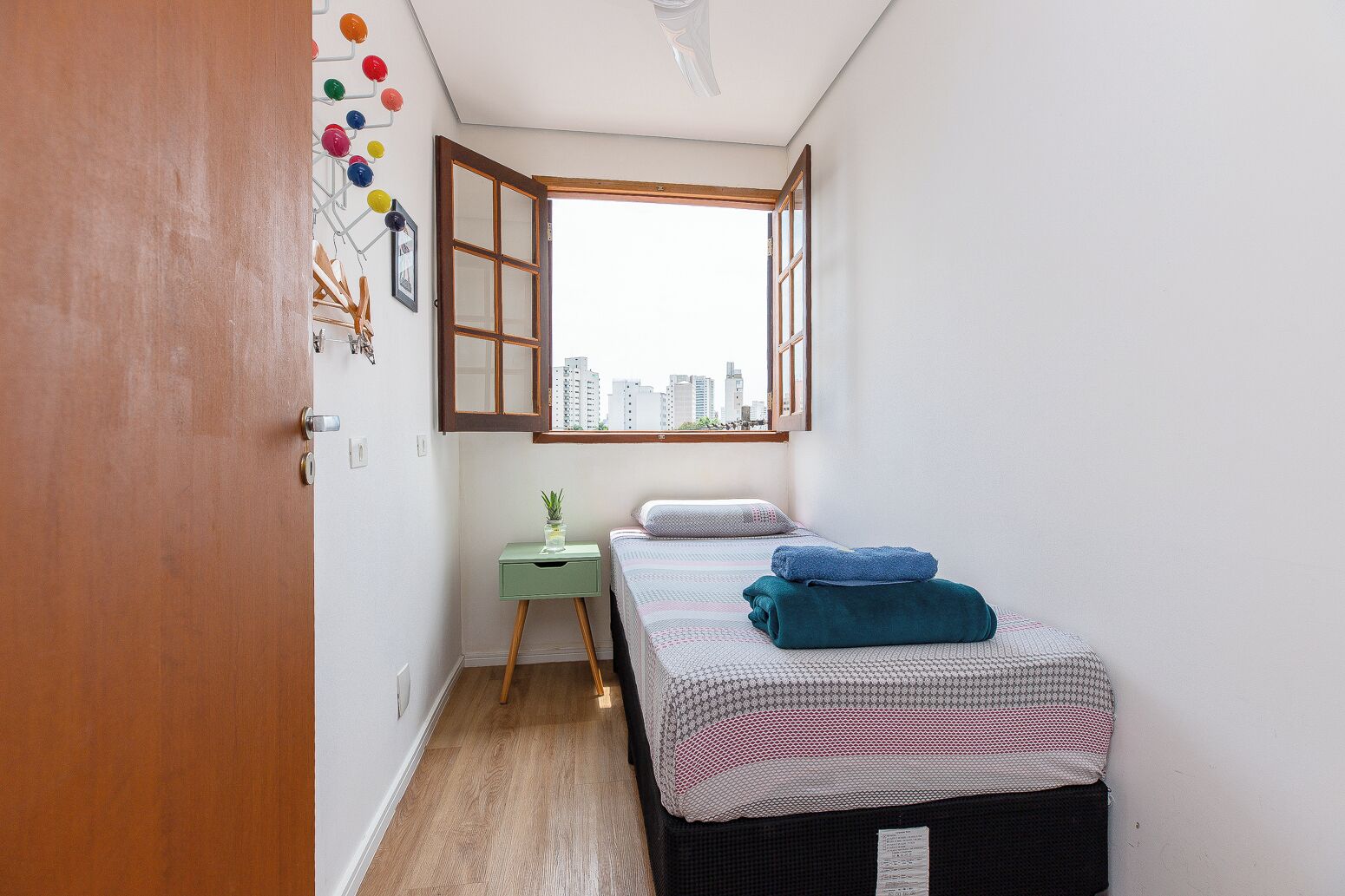 A small, cozy bedroom with a single bed, a green nightstand, a window with an open shutter, and a city view.