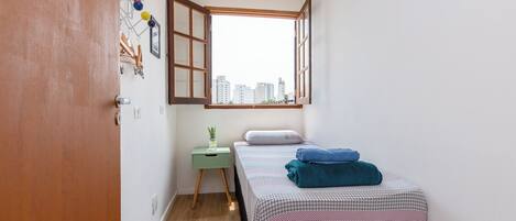 A small, cozy bedroom with a single bed, a green nightstand, a window with an open shutter, and a city view.