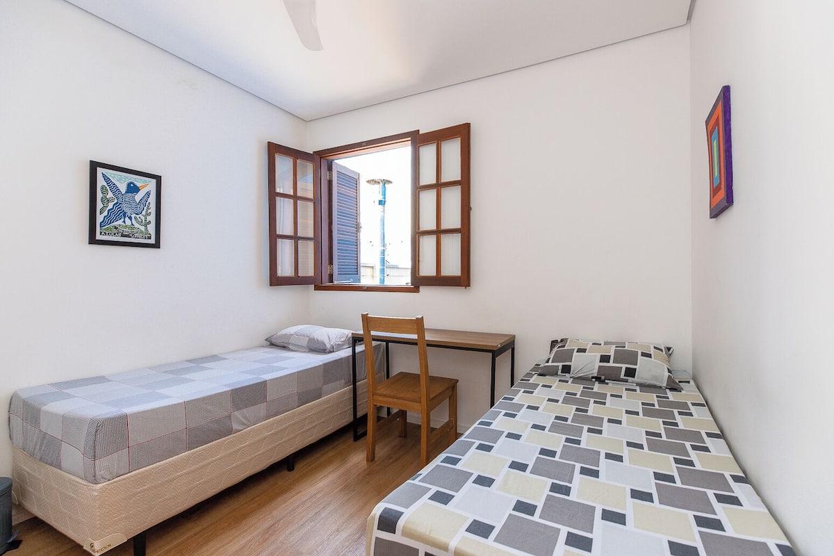 A small, well-lit room with two single beds, a wooden chair, a desk, and a window with open shutters.