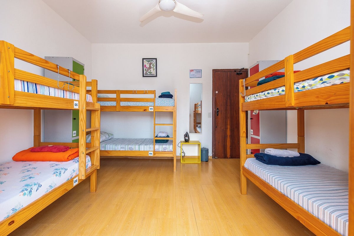 A dormitory room with bunk beds, wooden flooring, a ceiling fan, a small table, and a door.
