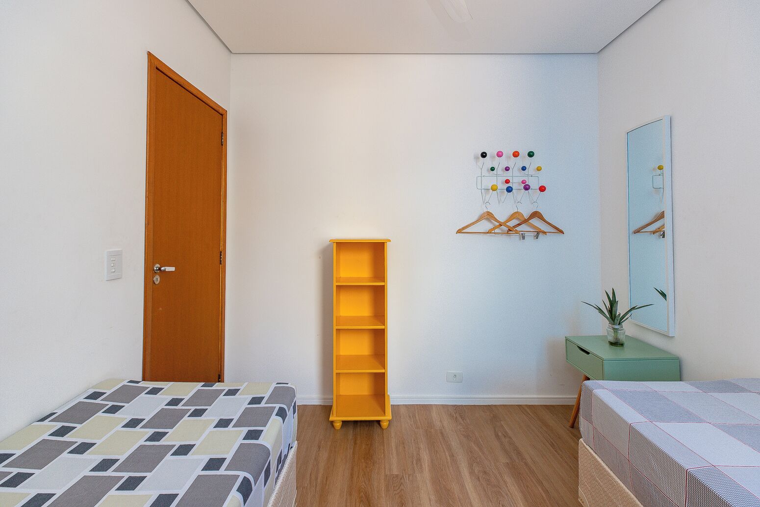 Ein kleines, ordentliches Schlafzimmer mit einer Holztür, einem Bett mit gemustertem Überwurf, einem gelben Regal, einem Wandtrockner, einem Spiegel und einer kleinen Pflanze auf einer grünen Kommode.