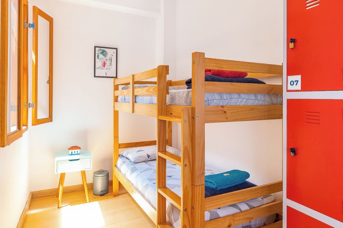 A dormitory room with bunk beds, a desk, a trash bin, and a red storage unit.