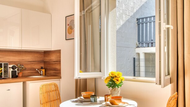 A compact kitchen with a white countertop, a small refrigerator, and a window with a view of a balcony. - La Suite Soleil - Maison studio avec jardin privé (Décines-Charpieu)