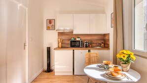 A compact kitchen with white cabinets, a microwave, and a small refrigerator. There is a dining table with a vase of sunflowers and a chair. - La Suite Soleil - Maison studio avec jardin privé (Décines-Charpieu)