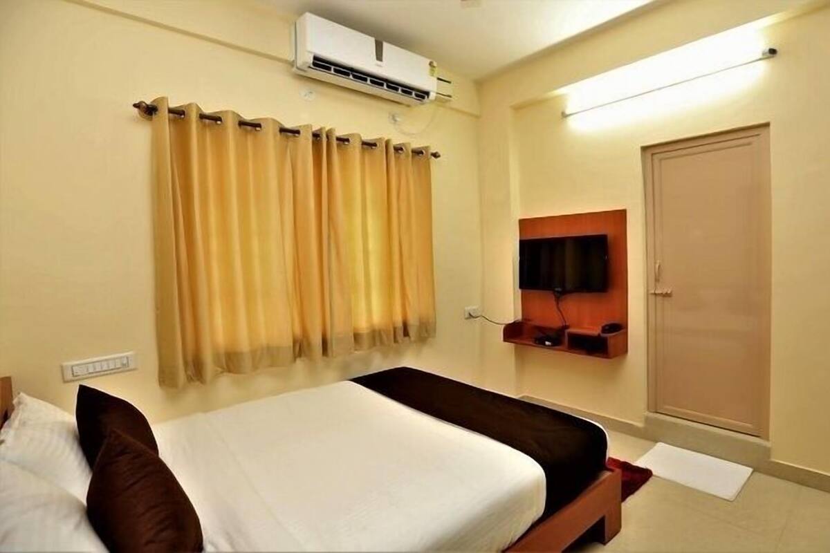 A hotel room with a bed, a TV mounted on the wall, an air conditioning unit above the bed, and a door.