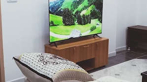 Olohuoneessa on televisio puisen jalustan päällä, kuviollisia koristetyynyjä sisältävä sohva ja laatoitettu lattia.